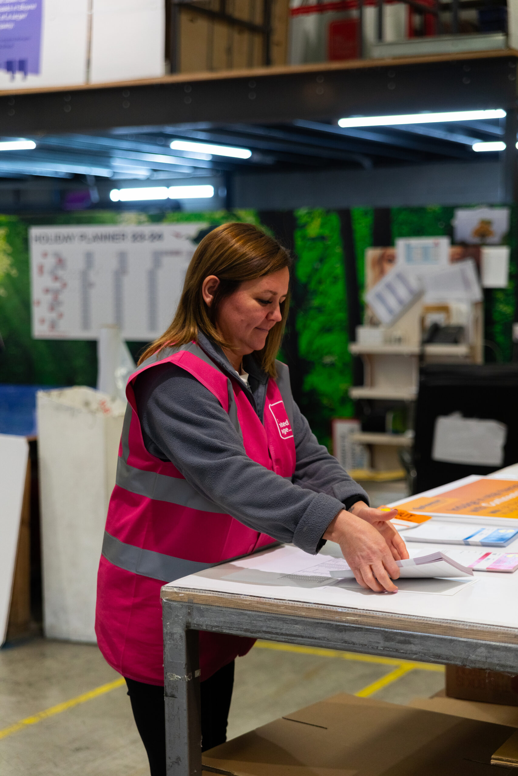 woman wearing a pink high vis vest with brown hair holding a stack of pages